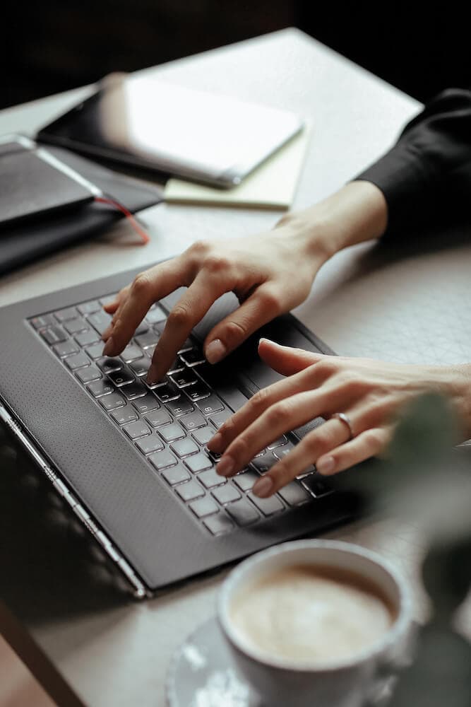 Close-up of a person typing on a laptop with a notebook and coffee cup on the desk.