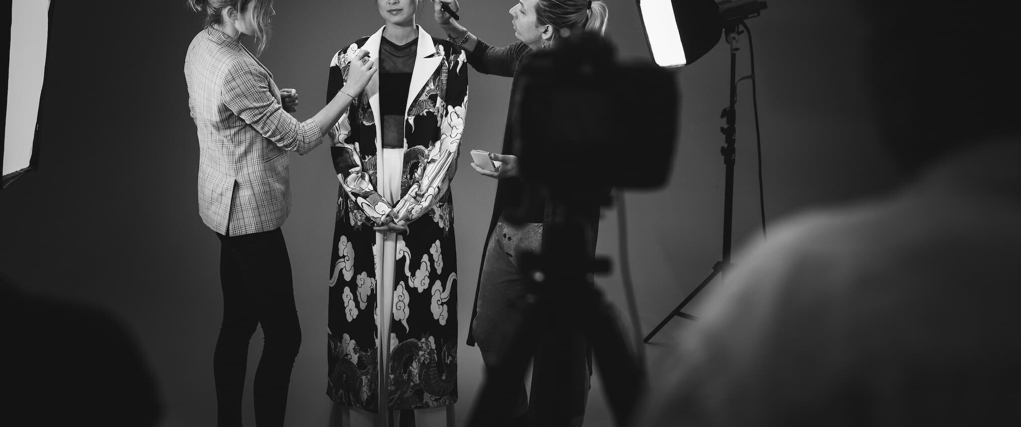 Behind the scenes of a photoshoot, with two makeup artists working on a model in a studio setting.